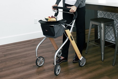 A woman in a kitchen pushing a rollator that has a plate of food on it. 