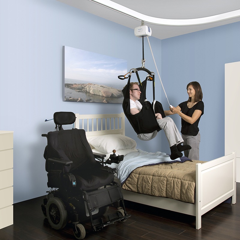 A woman helping a man get into bed using a ceiling lift.