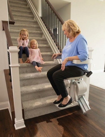 A grandmother using a stair lift with two grandchildren sitting on the steps waiting for her.