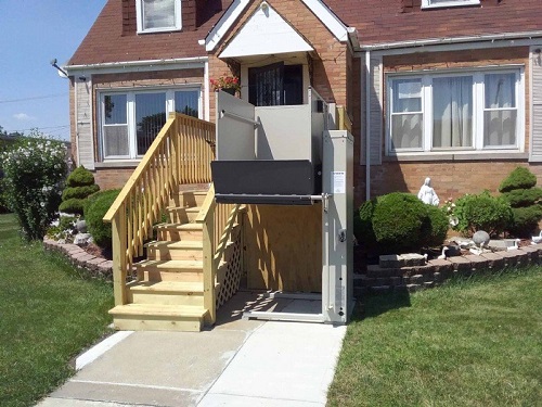 A vertical platform lift installed next to a flight of steps that lead up to the front door of a home.
