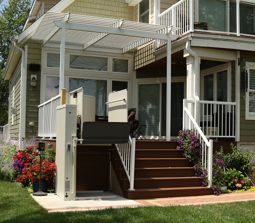 A wheelchair lift installed next to a flight of outdoor steps that lead to a large porch covered by a pergola.